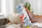 Preview: Thermobecher mit einem kunstvollen Weihnachtsstern-Design in Rot- und Blautönen, gehalten in einer Hand vor einem gemütlich beleuchteten Innenraum mit unscharfen, warmen Lichtakzenten im Hintergrund.